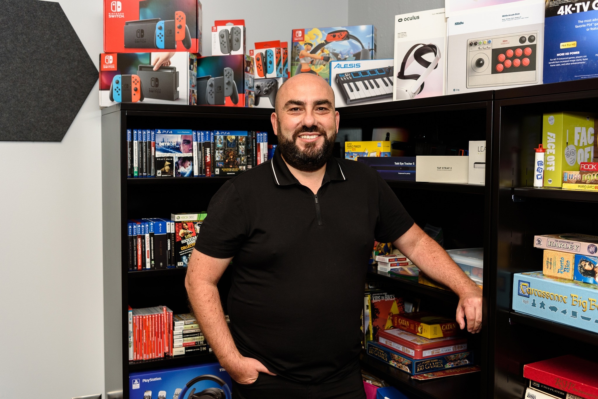 Dr. Şengün in front of a library of board and digital games and game consoles.